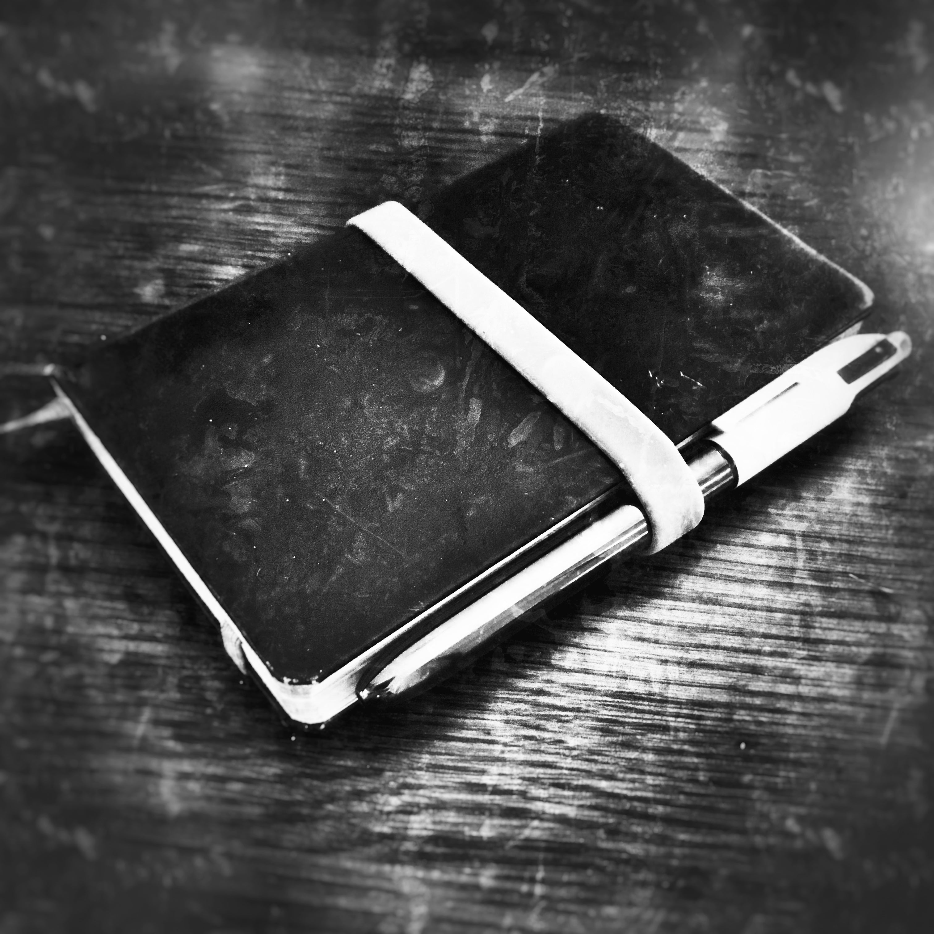 A black hardcover notebook with a white elastic closure band and a silver pen resting inside, placed on a dark textured wooden surface.